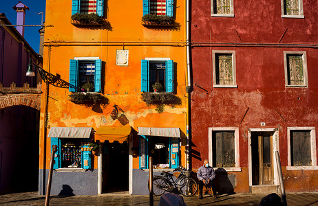 意大利老人坐在自家门外的自行车旁边，位于布拉诺岛色彩缤纷的房屋后面