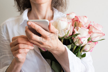 身穿白衬衫的中年妇女拿着智能手机和粉色和奶油色的玫瑰（裁剪和选择性聚焦）