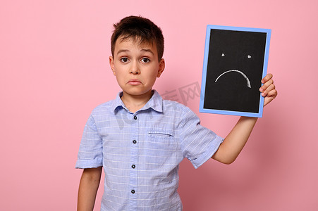 一个穿着蓝色衬衫的沮丧男孩在他的脸附近拿着一块黑板，上面画着表达沮丧、悲伤和失望的彩绘。