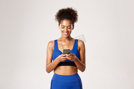 身着蓝色运动服、使用手机、站在白色背景下的健康而健康的非洲裔美国女运动员