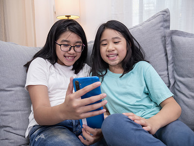 坐在客厅沙发上的快乐兄弟姐妹一起用手机与父母交谈，微笑的大女孩通过智能手机显示可爱的小妹妹视频通话与家人交谈。