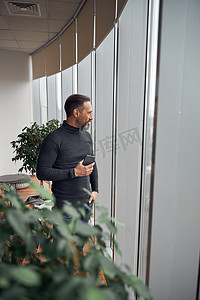 快乐聪明的成年白人商人站在办公空间的大窗户附近，手里拿着智能手机