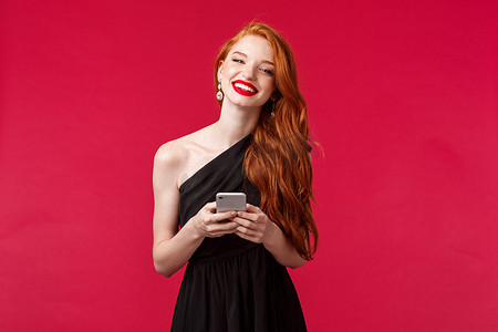 身着优雅黑色舞会礼服、留着姜黄色头发、手持智能手机、发短信、使用应用程序、想要拍照、笑着看相机、红色背景的华丽微笑女性肖像