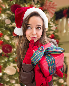 思考的女孩戴着圣诞圣诞老人帽子，在装饰树前用蝴蝶结包裹礼物。