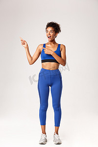 穿着蓝色运动服的非洲裔美国女运动员全长惊讶，手指向左，惊奇地看着，展示了一些关于锻炼和健身房的东西，白色背景