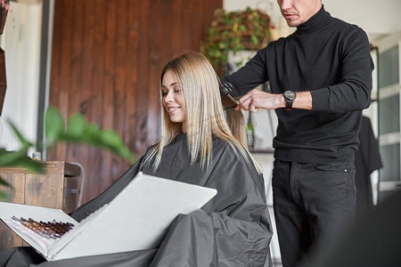 发型女摄影照片_金发女客户正在选择书中的颜色，而美发师正在剪头发
