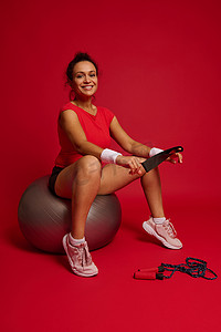 跳绳动态摄影照片_迷人迷人的西班牙裔女运动员拿着弹性健身带，在红色背景下锻炼后坐在健身球上。