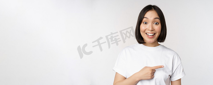 手指向右摄影照片_快乐美丽的亚洲女孩在空白的复制空间上展示广告，手指向右，微笑，展示横幅或公司标志，白色背景