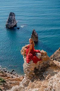 飘逸长裙摄影照片_一位身穿红色长裙、头发飘逸的女孩坐在海面上的岩石上。