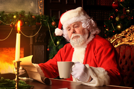 他在家摄影照片_快乐圣诞老人的画像坐在他在家里靠近圣诞树的房间里，阅读圣诞信或愿望清单。