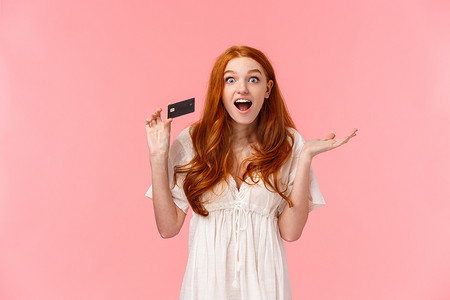 上腰画像被逗乐、惊奇和兴奋的时尚女性红发女孩讲述银行里的大促销和奖金，拿着信用卡，微笑着惊讶的粉红色背景
