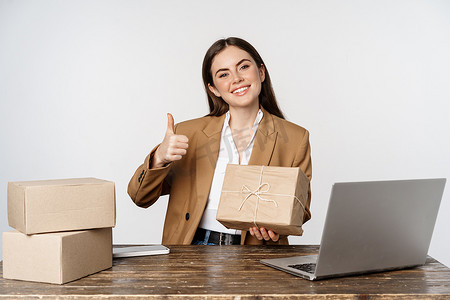 微笑的女商人展示包裹、盒子里的订单和竖起大拇指，在自己的商业商店工作，坐在笔记本电脑旁，白色背景