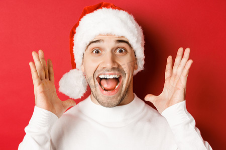 戴着圣诞帽的快乐年轻人的特写，发布重大的圣诞公告，微笑着惊讶，站在红色背景上