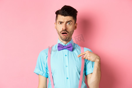 穿着领结和吊带裤的搞笑搞笑男子的形象，指着自己，好像被指控或被选中，惊讶地扬起眉毛，站在粉红色的背景上
