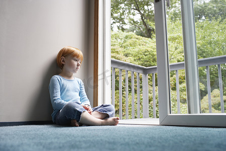 全长沉思的年轻女孩坐在家里的地毯上，透过阳台望去