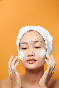 年轻的亚洲女性在脸上涂抹奶油，并戴着浴巾触摸脸部。