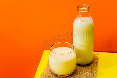 玻璃瓶和大玻璃杯加牛奶，放在红色背景的黄色桌子上的质朴织物正方形上。