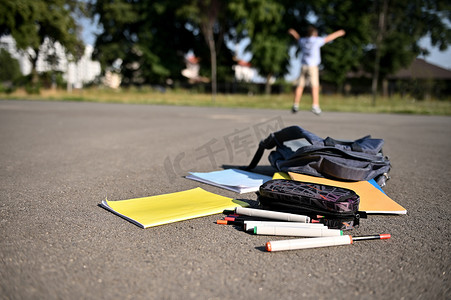 散落的学习用品和作业本从一个打开的背包里掉出来，躺在校园的柏油路上，背景是一个模糊的快乐男生举起双手