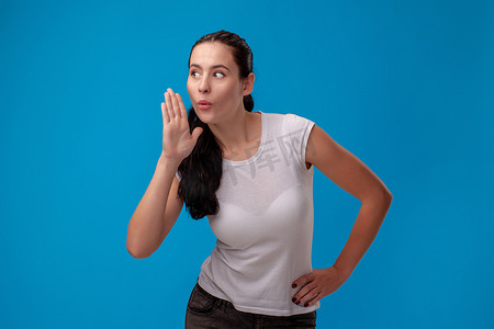 一位身穿白色 T 恤的年轻漂亮女子在蓝色墙壁背景下的工作室肖像。