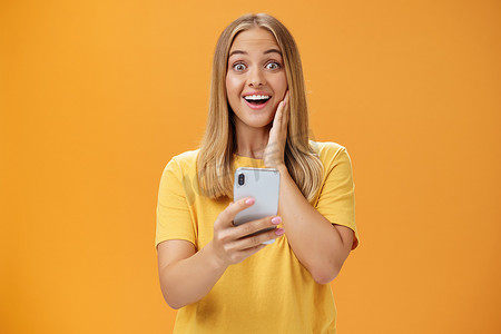令人惊讶和印象深刻的女性对智能手机中令人敬畏的应用程序做出反应的肖像，惊讶和喜悦地对着相机手持手机在橙色墙壁上大笑