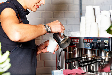 空包纸杯摄影照片_一位身穿黑色围裙的咖啡师在咖啡机的背景下将新鲜准备的咖啡倒入纸杯中，空纸杯的幻灯片模糊不清。