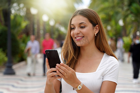 兴奋的年轻女子在街上行走时看着她的智能手机，背景上的人模糊。