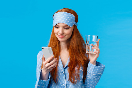 上腰肖像快乐的红发欧洲魅力女孩戴着睡眠面罩和睡衣，拿着玻璃水并检查饮用计划应用程序，微笑着通过智能手机聊天，蓝色背景