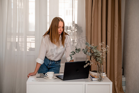 欧洲职业女性坐在家庭办公室的桌子旁，手里拿着一台笔记本电脑，一位积极的女性一边在 PC 上工作一边学习。