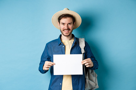 快乐的游客背着背包，展示着一张空纸，对着镜头微笑，站在蓝色背景上。