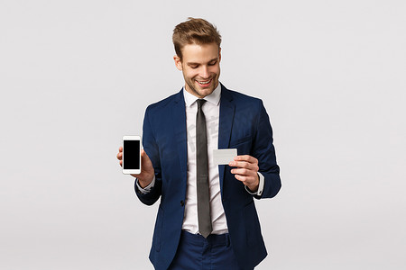 自信的年轻金发商人，身穿蓝色经典西装，手持智能手机和信用卡，展示移动显示、在线支付方法、财务应用、站立白色背景