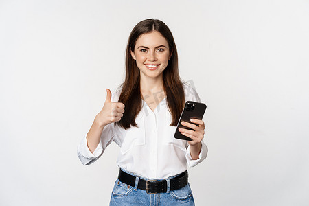 笑得开心的企业女性，女模特竖起大拇指，手持智能手机，使用手机，站在白色背景上