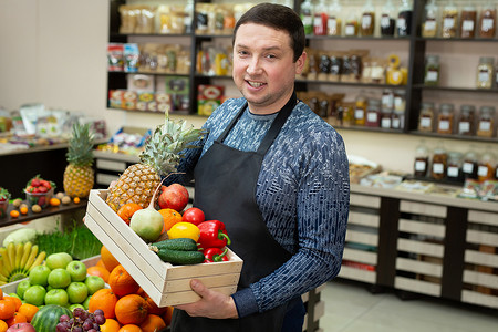 微笑的男推销员在店里拿着一个木箱，里面装着蔬菜和水果