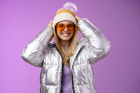 下雪保暖摄影照片_精力充沛、大胆时髦的年轻迷人女性，在冬季旅行中与朋友玩得开心，学习单板滑雪，面带微笑，享受假期，戴上银色保暖夹克太阳镜，紫色背景