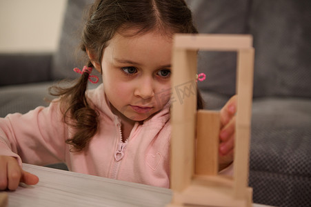 粉色木块摄影照片_可爱孩子的特写肖像，穿着粉色运动衫的学龄前女孩专注于用木块建造木结构。