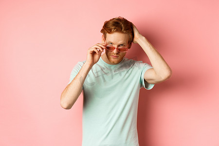 戴着墨镜的英俊年轻红发男子，用手梳理头发，站在粉红色背景上，对着镜头显得自鸣得意和自信