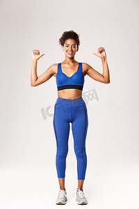 身穿蓝色运动服、自信满满的非洲裔美国女运动员，带着自豪而快乐的微笑指着自己，展示锻炼进展，站在白色背景上