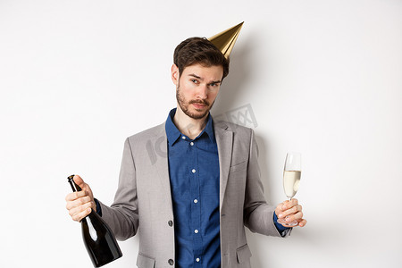 派对男戴着生日帽庆祝，手里拿着香槟酒瓶和玻璃杯，看起来喝醉了，穿着灰色西装