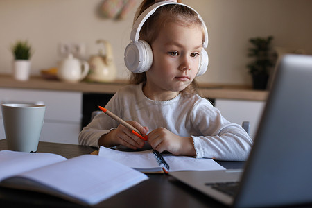 初等教育摄影照片_戴着耳机的微笑小女孩在家里使用笔记本电脑在线学习手写，戴着耳机的可爱快乐的小孩在 PC 上上互联网课程或课程。
