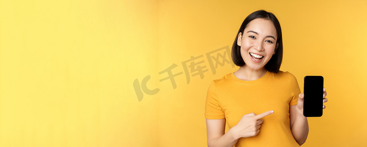 美丽快乐的亚洲女孩展示手机屏幕，在智能手机小工具上的应用，站在黄色背景上