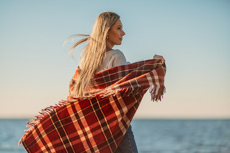 迷人的金发白种女人在日落时分在海滩上享受时光，裹着毯子走着，看着旁边，背景是落日的天空和大海。