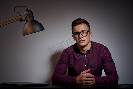戴着眼镜和衬衫的商人坐在黑暗房间的桌子旁，房间里有一盏灯