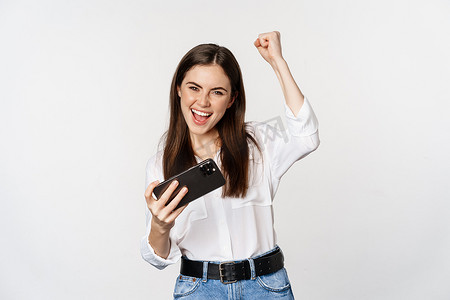 快乐的黑发女人玩手机视频游戏，微笑着看着屏幕兴奋，站在白色背景上