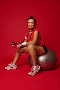 穿着红色紧身上衣和黑色运动短裤的漂亮运动女性拿着跳绳，对着镜头微笑，同时坐在红色背景的银色健身球上，有复制空间