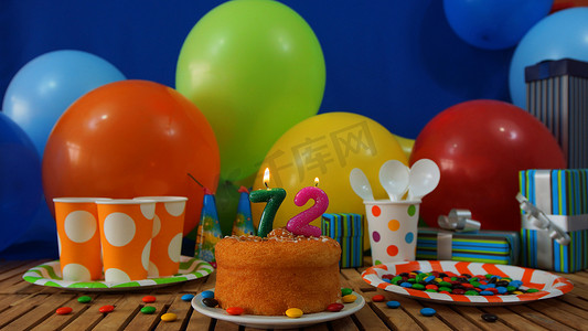 气球墙摄影照片_质朴木桌上的生日蛋糕，背景是彩色气球、礼物、塑料杯和塑料板，背景是糖果和蓝墙
