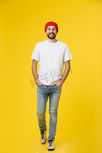 背带裤摄影照片_年轻的时髦男人戴着帽子，背带裤，在黄色背景上隔离。