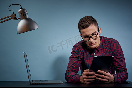 一个戴眼镜的男人坐在办公室的桌子旁，那里有一台笔记本电脑和一盏台灯