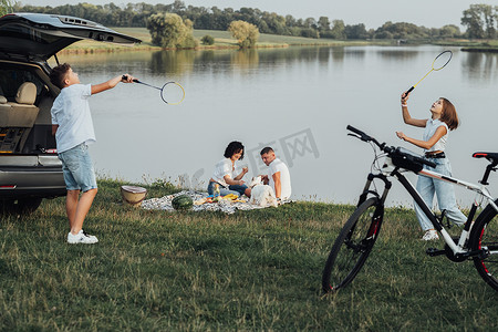 快乐的四口之家与宠物狗在户外野餐，两个十几岁的孩子打羽毛球，而父母坐在湖边