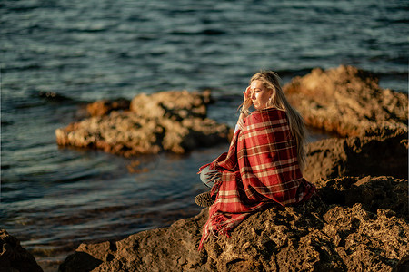 迷人的金发白种女人在日落时分在海滩上享受时光，坐在毯子里，看着一边，背景是落日的天空和大海。