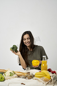 身穿绿色 T 恤的快乐女人打开购物网状生态袋，里面装着健康的纯素蔬菜、水果、面包、零食，手里拿着西兰花和黄胡椒。