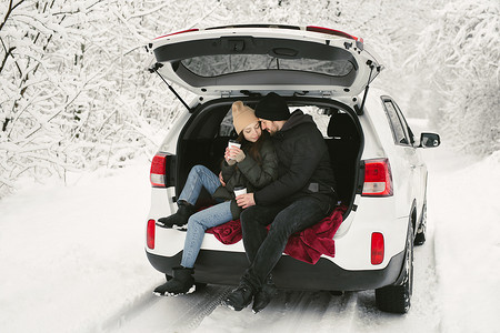 一对年轻夫妇，一男一女，在冬天、白雪皑皑的森林里坐在汽车后备箱里，拥抱、亲吻和喝咖啡